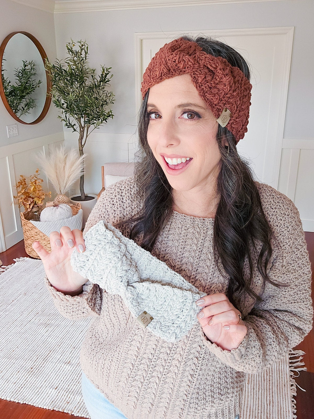 A woman in a knit sweater and a headwrap smiles while holding a light-colored knitted headwrap in a well-lit room with neutral decor, surrounded by vintage clothes and unique jewellery.