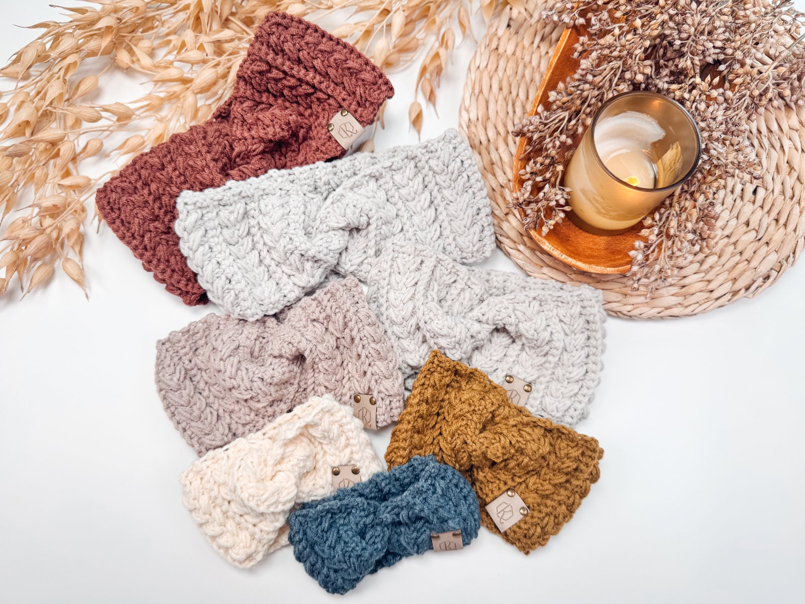 A collection of knitted headbands in various colors arranged on a white surface with dried flowers and a lit candle placed on a woven mat, creating the perfect setting for a handmade gift.