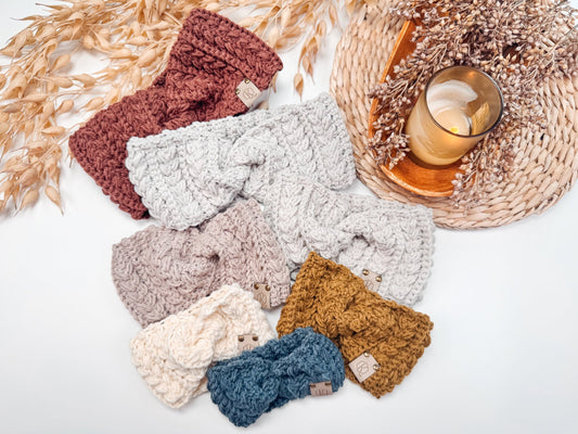 A collection of knitted headbands in various colors arranged on a white surface with dried flowers and a lit candle placed on a woven mat, creating the perfect setting for a handmade gift.
