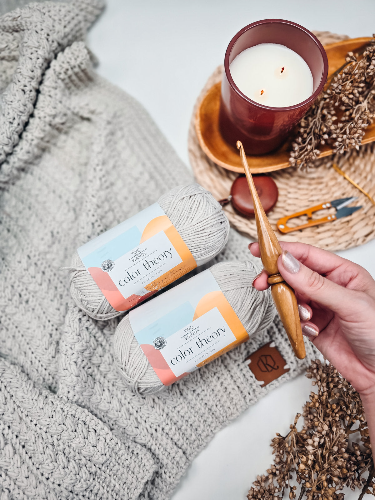A hand holding a wooden crochet hook over a gray crocheted fabric, with two balls of light gray yarn labeled "color theory," a lit candle, and a pair of scissors on a woven tray adorned with dried flowers next to a textured crochet throw blanket.