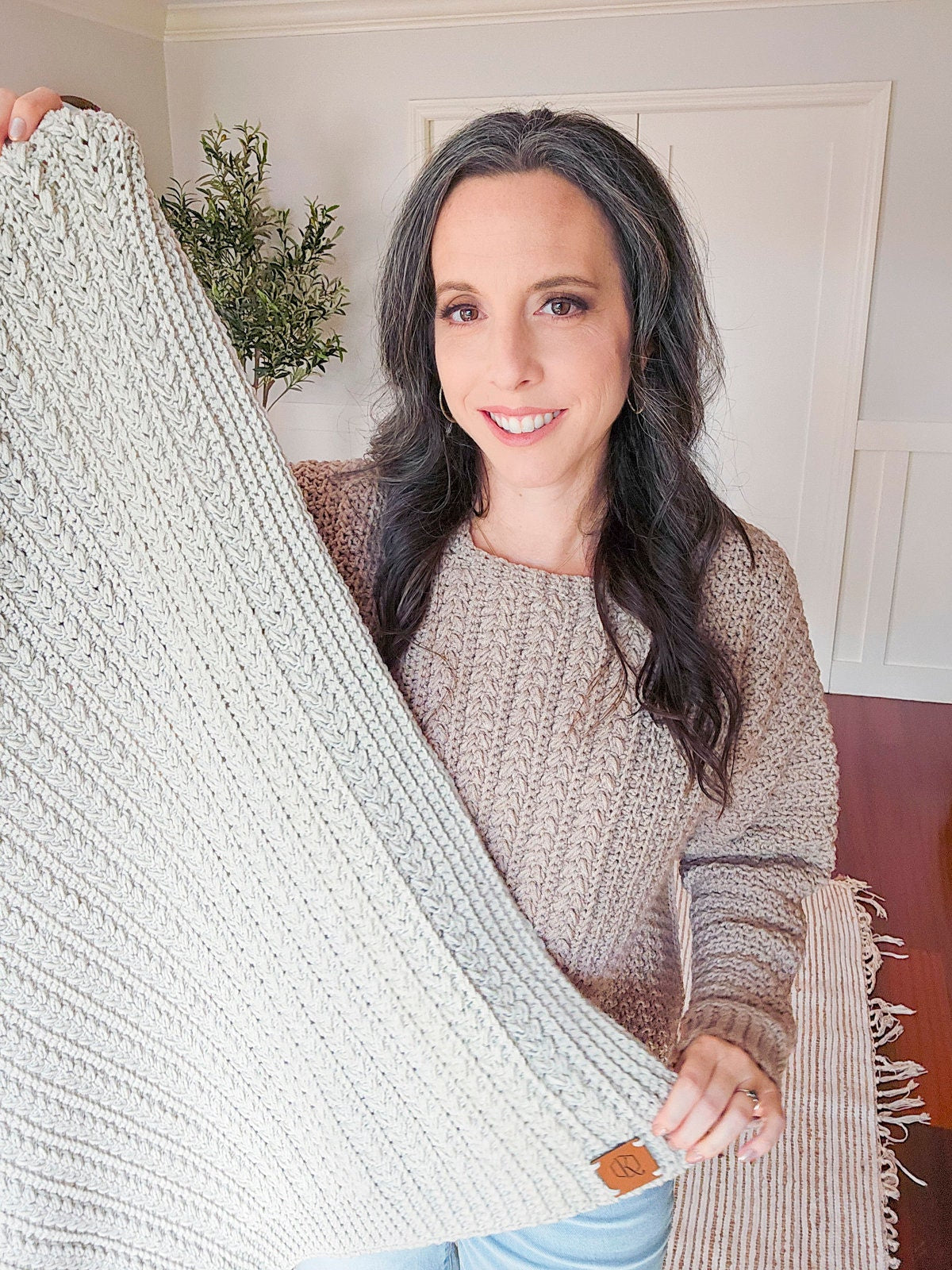 Person holding a textured, light-colored crochet blanket, standing in a room with a plant in the background. The cozy wrap was a thoughtful handmade gift perfect for chilly evenings.