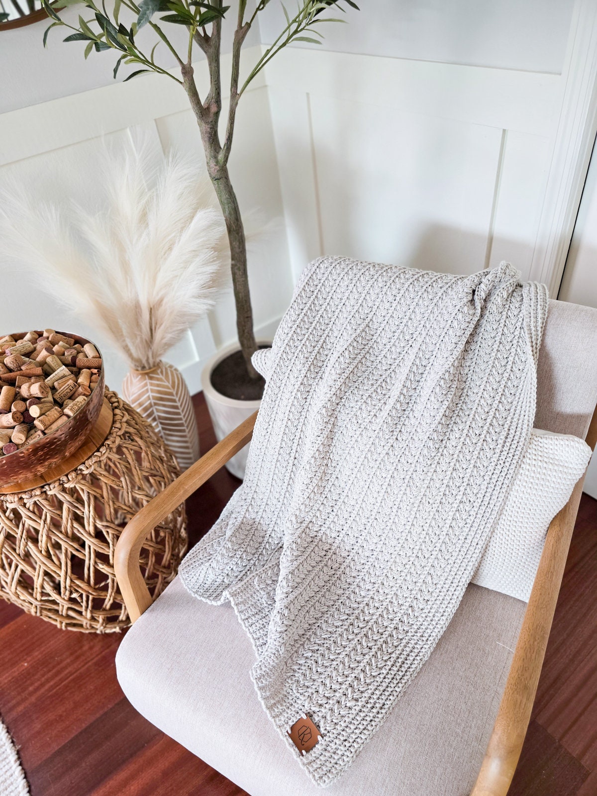 A cream-colored crochet blanket is draped over a beige chair next to a potted plant and a wicker side table with a bowl of corks, creating the perfect nook for showcasing a crochet blanket.