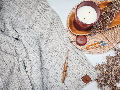 Light gray crochet blanket laid flat with a wooden crochet hook, candle, snips, and dried flowers on a woven mat