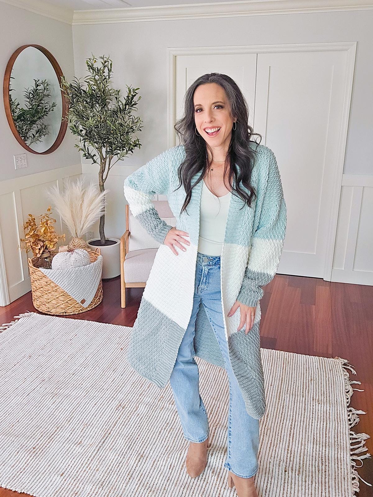 A woman stands in a living room wearing a color-blocked cardigan, jeans, and boots. Adorning her outfit is unique jewelry that catches the light. The room features hardwood floors, a round mirror, and a decorative plant.