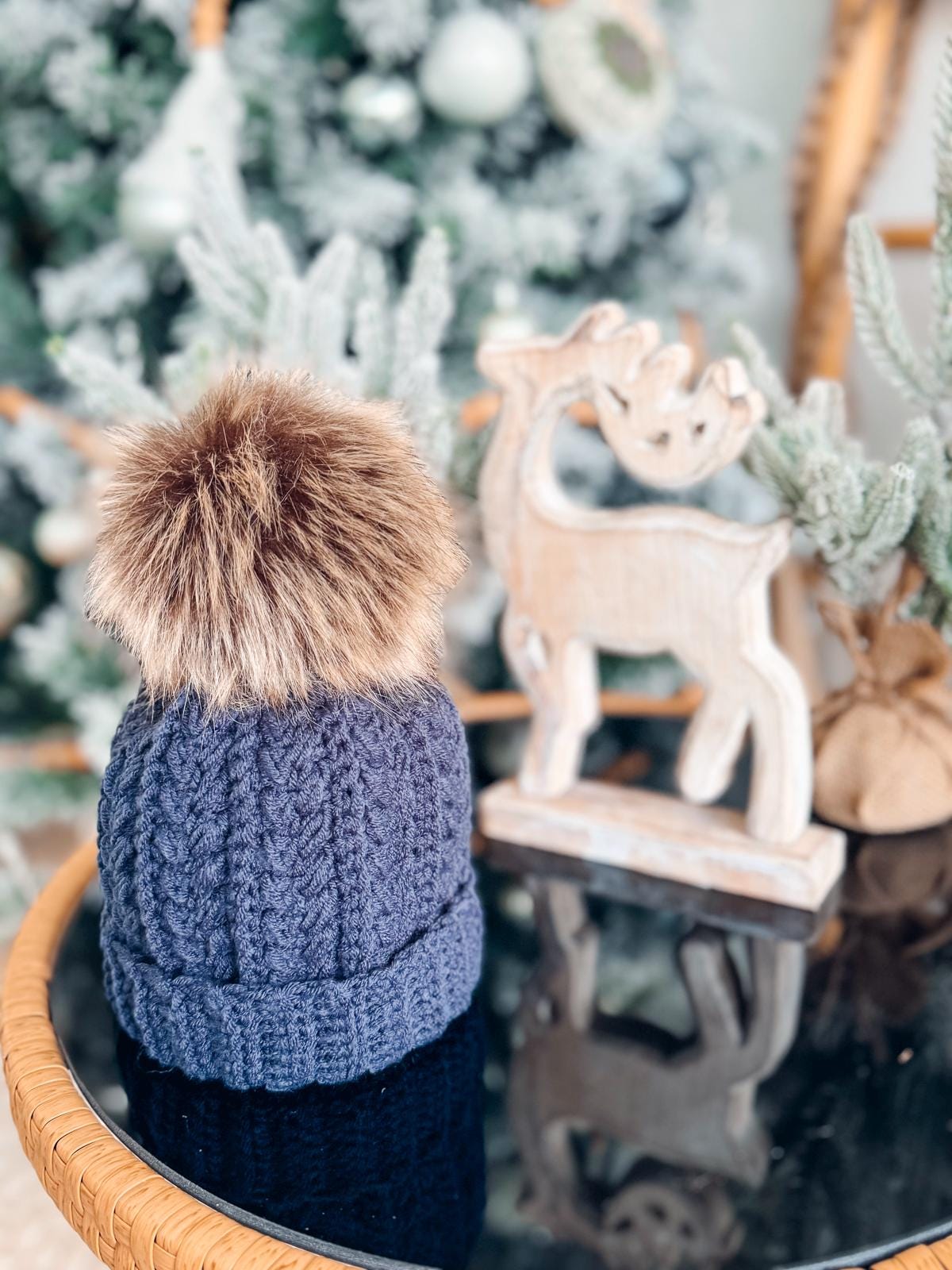 A crocheted blue hat with a faux fur pom-pom, resembling a handmade gift, sits on a glass table next to a wooden reindeer decoration. A decorated Christmas tree glows softly in the blurred background.