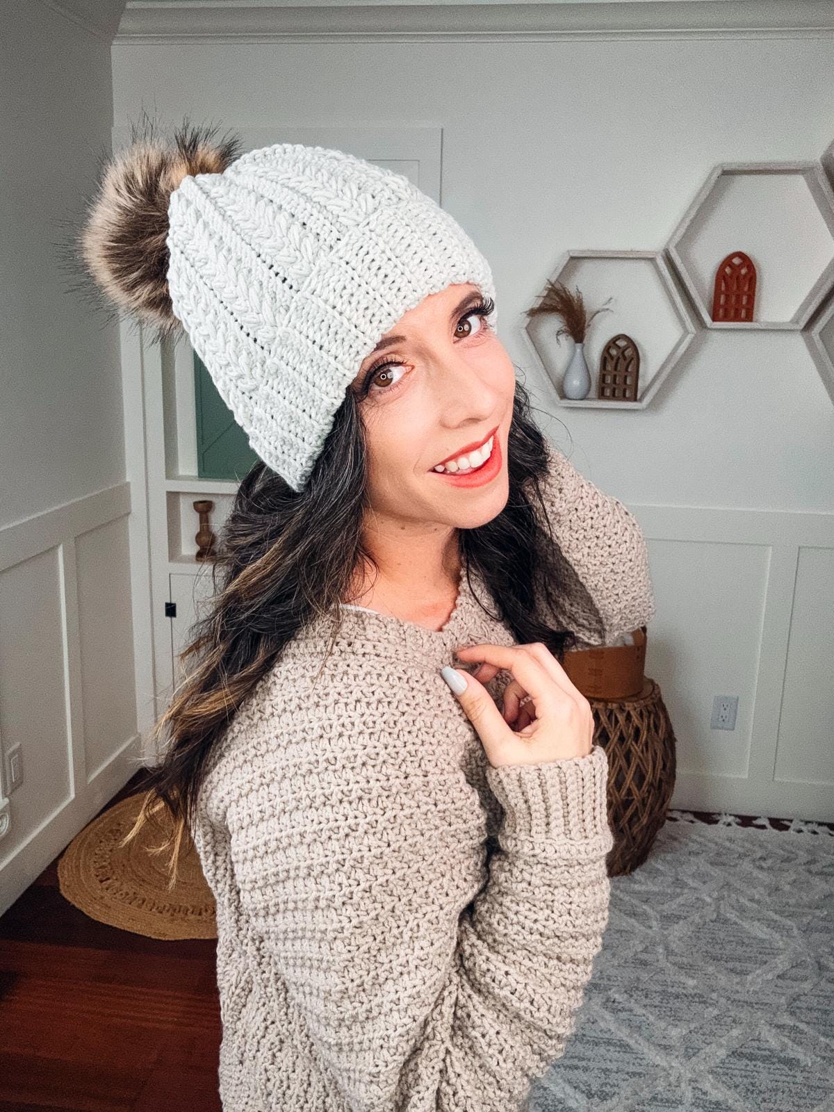 A woman in a crochet sweater and beanie stands indoors, smiling at the camera, adorned with unique jewellery.