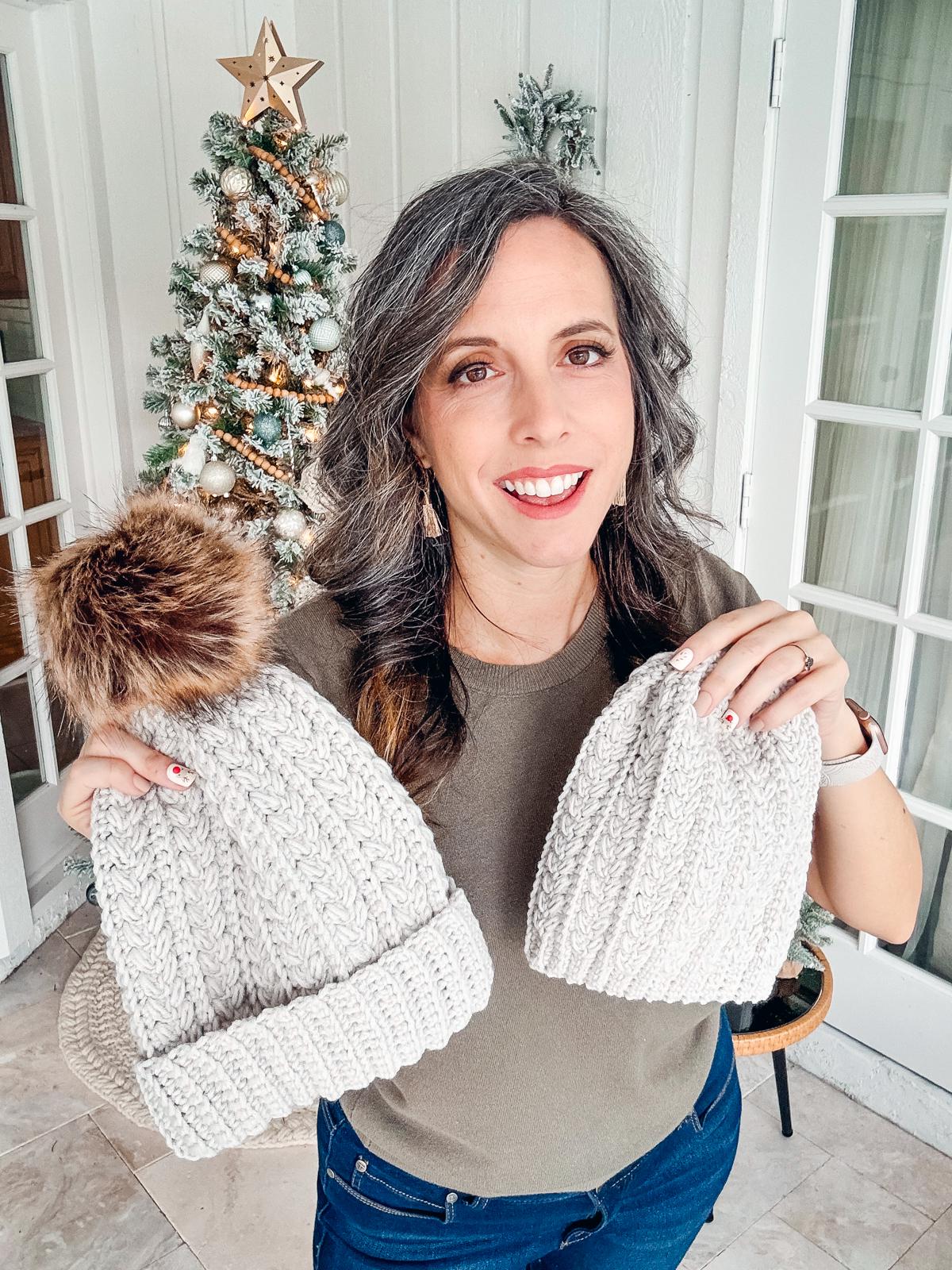 Woman holding two crocheted hats, standing in front of a decorated Christmas tree, each piece a handmade gift brimming with care.
