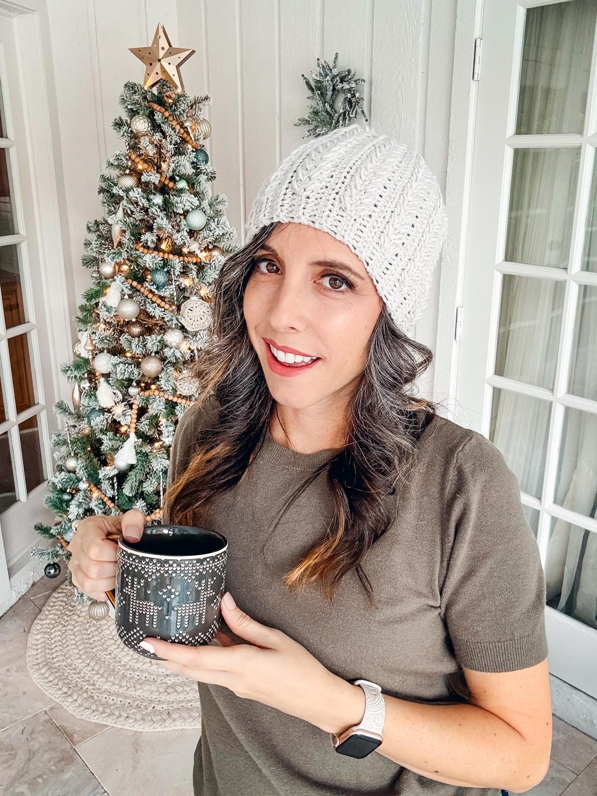 A woman in a crochet hat, wearing vintage clothes, holds a mug in front of a decorated Christmas tree.