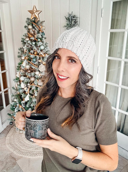 A woman in a crochet hat, wearing vintage clothes, holds a mug in front of a decorated Christmas tree.