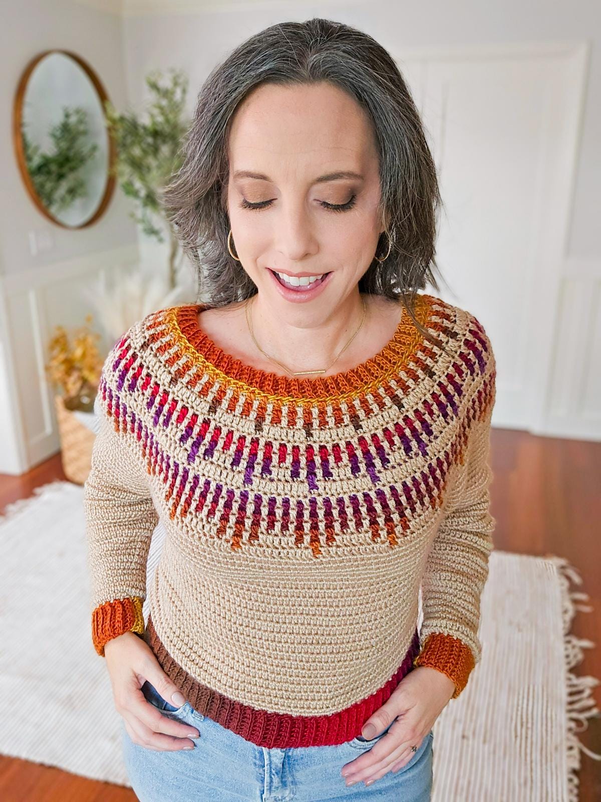 A person wearing a beige and multicolored crochet sweater, reminiscent of vintage clothes, stands indoors with eyes closed and a slight smile.
