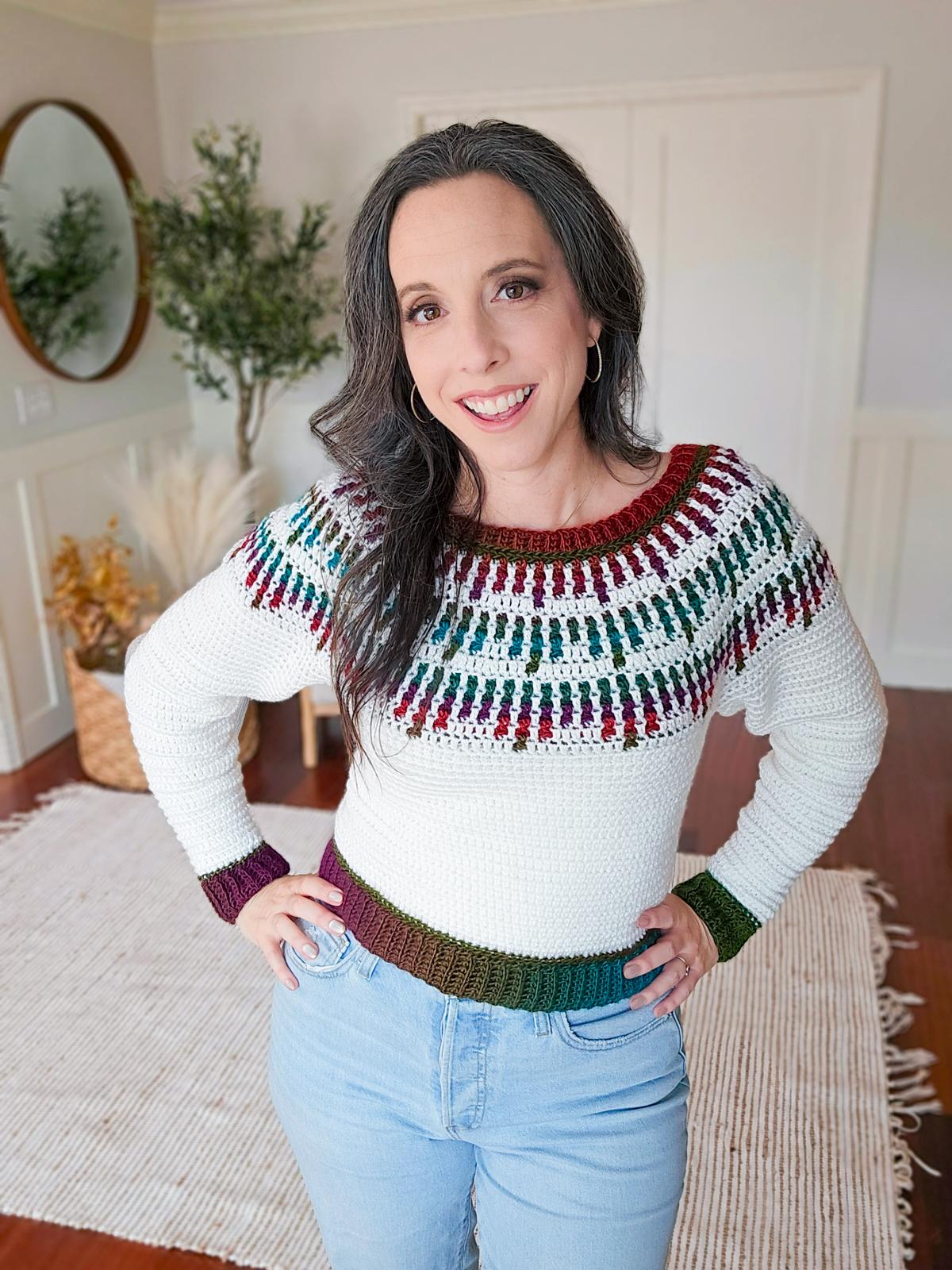 Person smiling, wearing a crocheted white sweater with a colorful patterned yoke and cuffs—a cherished handmade gift—standing in a room with a mirror, plant, and patterned rug.