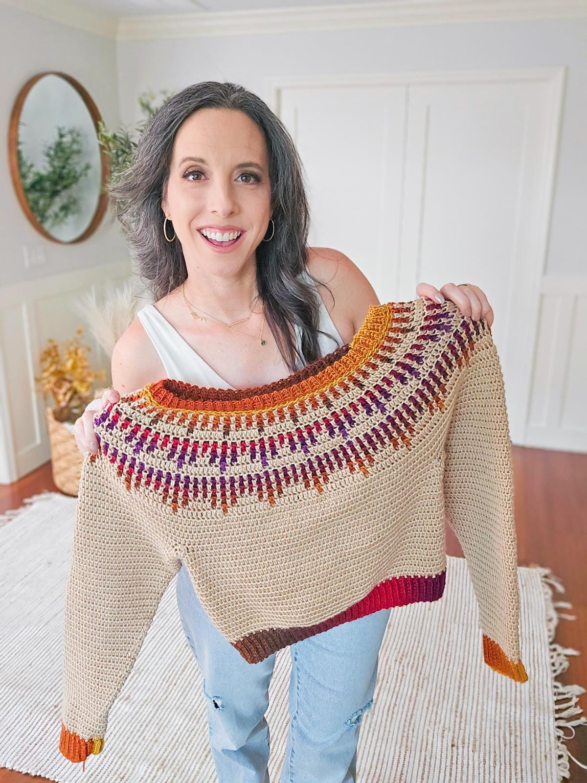 A woman smiling and holding a crochet sweater with a colorful mosaic yoke design, perfect as a handmade gift.