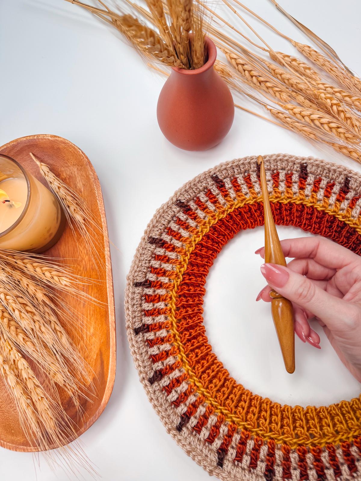 A hand holding a crochet hook works on a multicolored crocheted round placemat, crafting a charming handmade gift. Nearby are wheat stalks in a vase and tray, and a lit candle on the white surface, creating an inviting, cozy atmosphere.