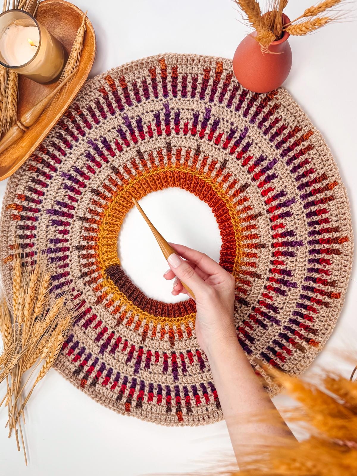 A hand holds a crochet needle, crafting a colorful, circular pattern. The decor includes a candle, vase, and wheat stalks—an inviting setting for creating crochet and the perfect handmade gift.