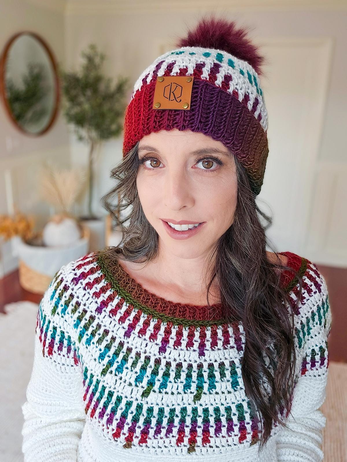 A person wearing a crocheted sweater and beanie with red and orange patterns stands smiling in a room adorned with a vintage mirror, alongside a plant.