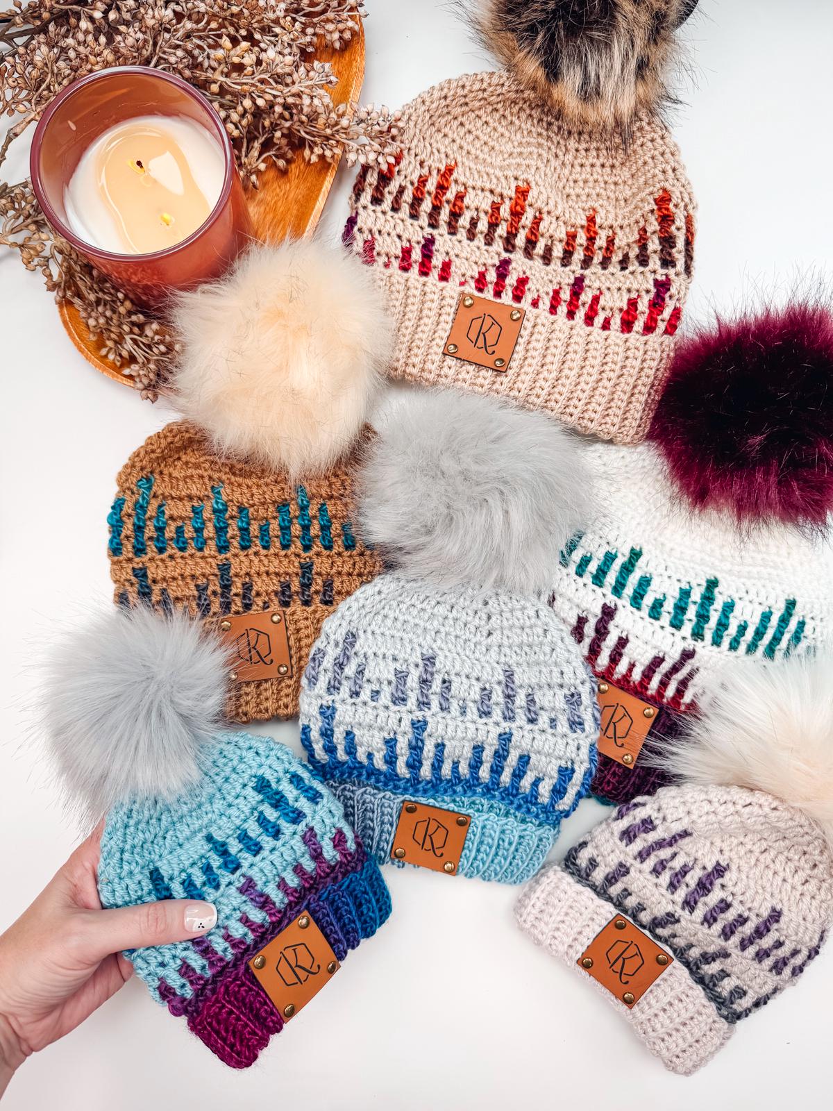 A collection of six knitted beanies with pom-poms, featuring various patterns and colors, arranged around a lit candle and decorative foliage. A hand is holding one of the beanies, offering it as a charming handmade gift.