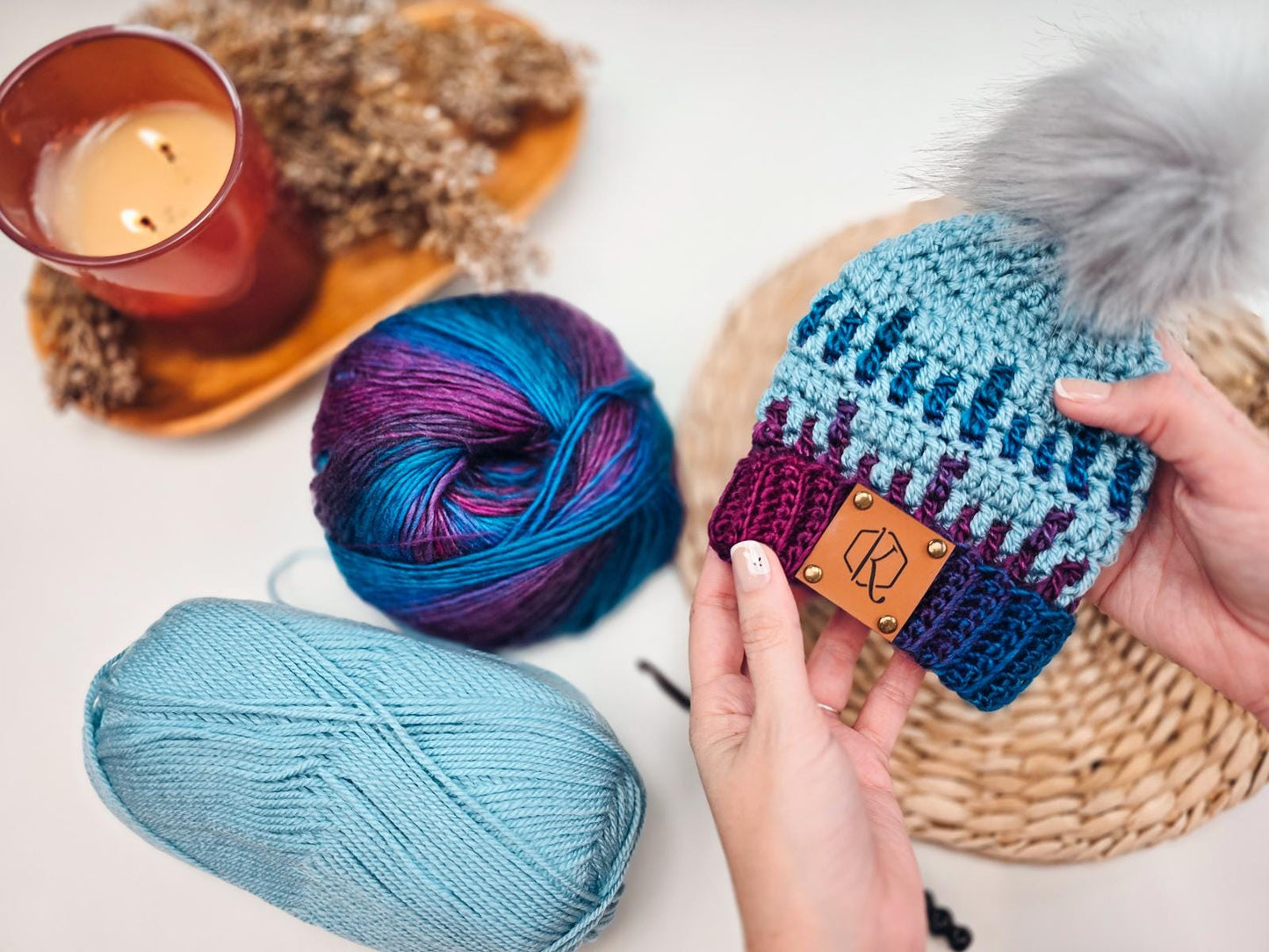 A person holds a knitted blue and purple baby hat with a pom-pom, surrounded by yarn balls, a red candle, and dried flowers—a perfect handmade gift for any occasion.