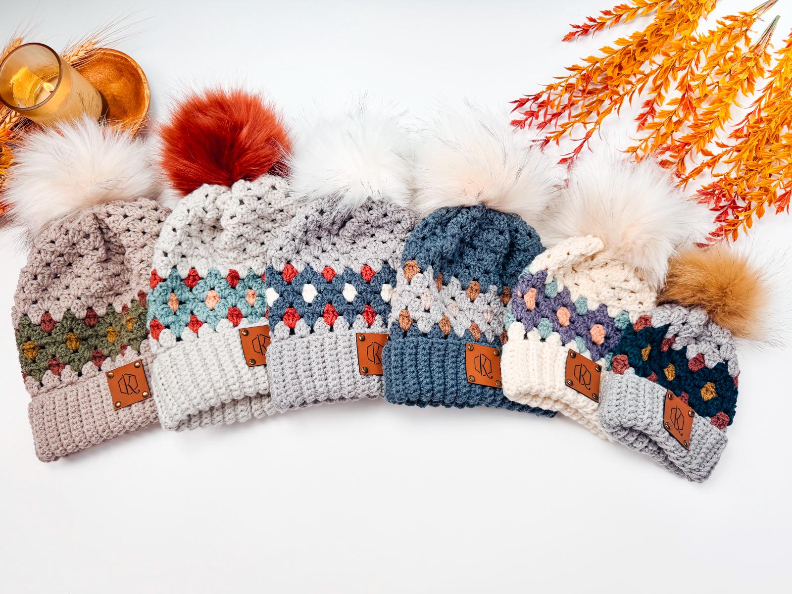 Five crocheted beanies with pom-poms in various colors are arranged in a row, accompanied by autumn leaves and a candle, creating the perfect handmade gift setting.