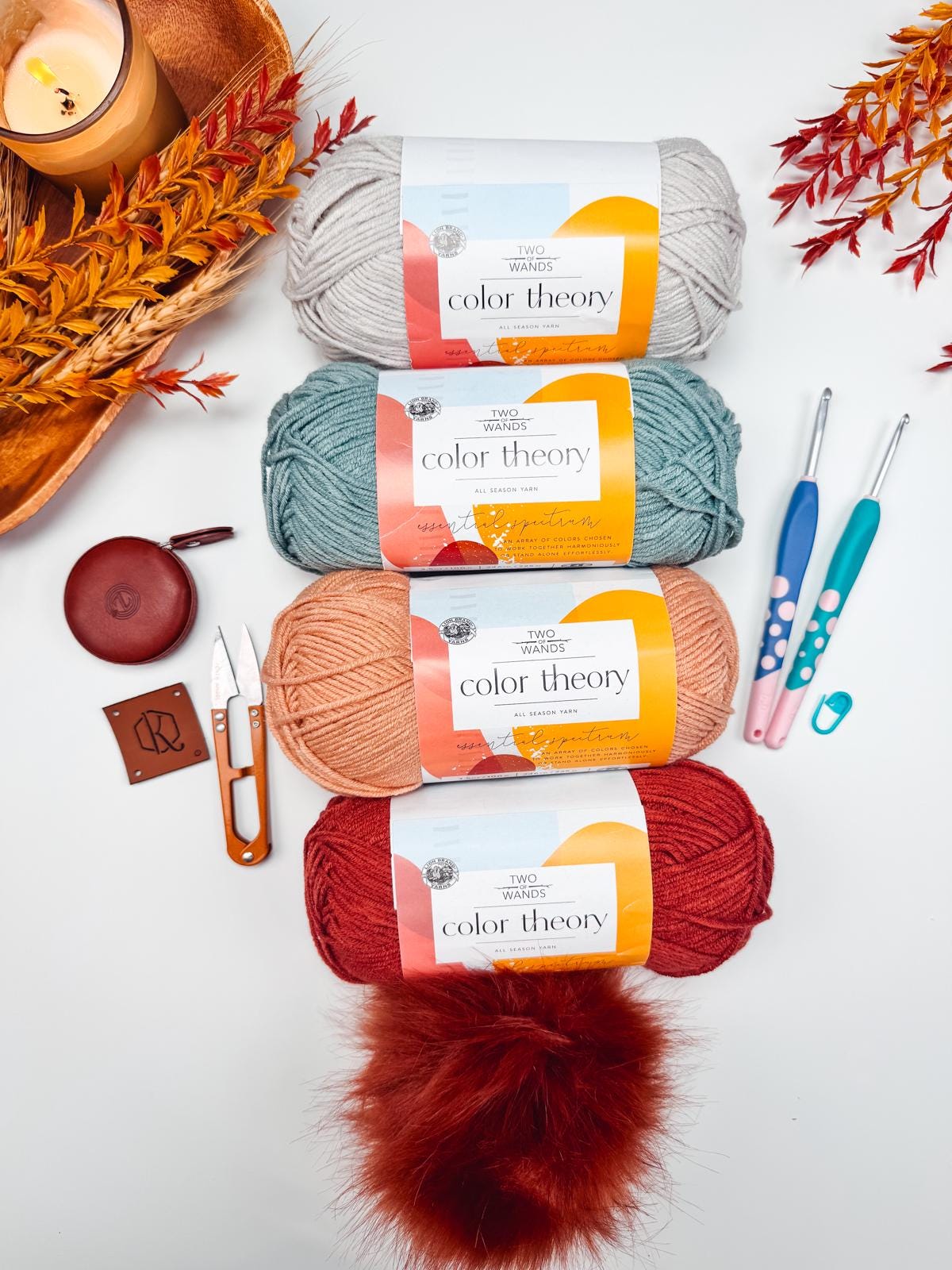 Yarn balls in gray, teal, peach, and red accompany crochet hooks, scissors, a candle, and red foliage on a white surface—perfect setting for crafting a handmade gift that's as warm and personal as it is beautiful.