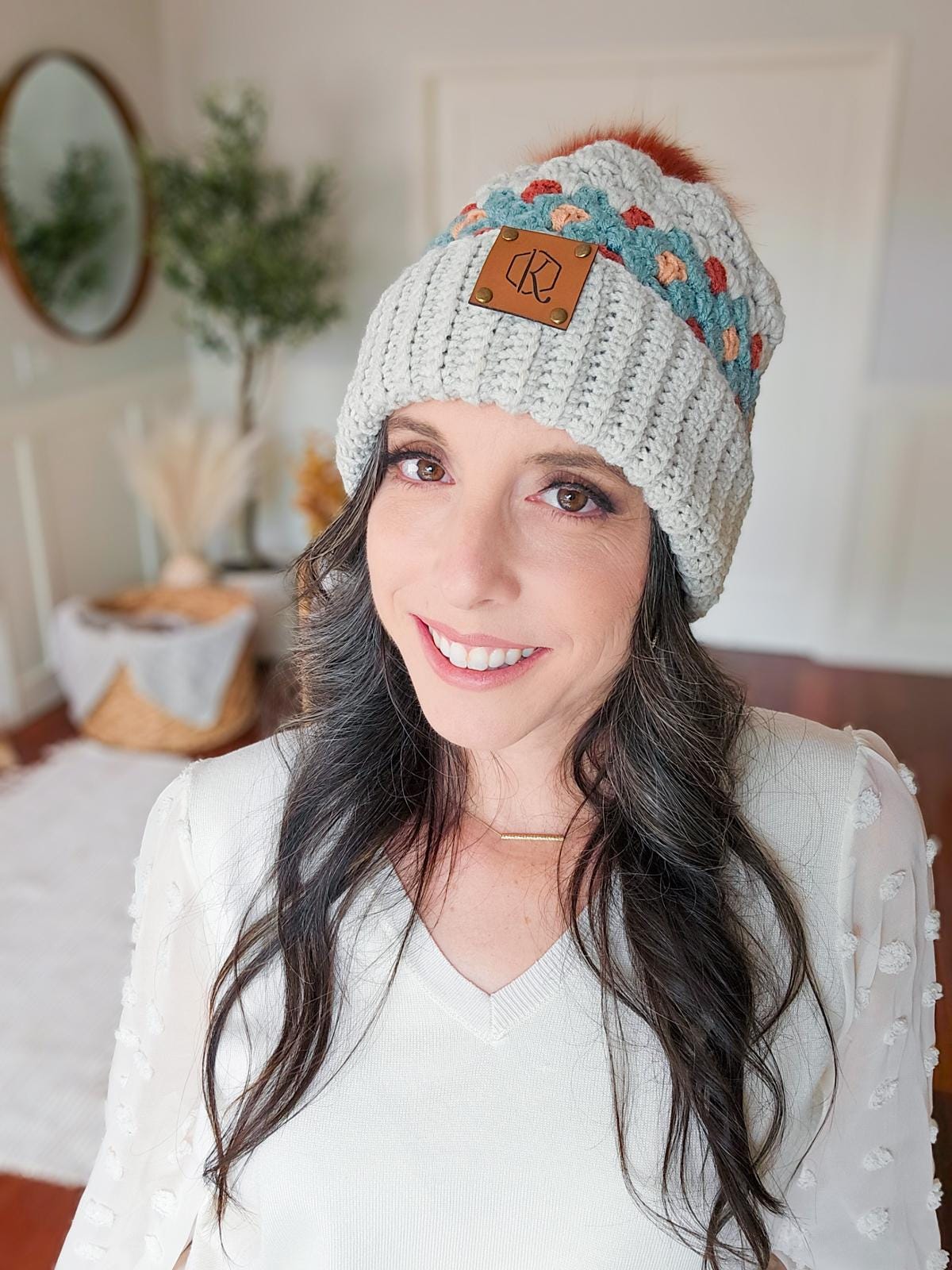 A woman smiles while wearing a knit hat with a colorful band and pom-pom. She stands indoors, surrounded by vintage clothes, with a plant and mirror in the background.