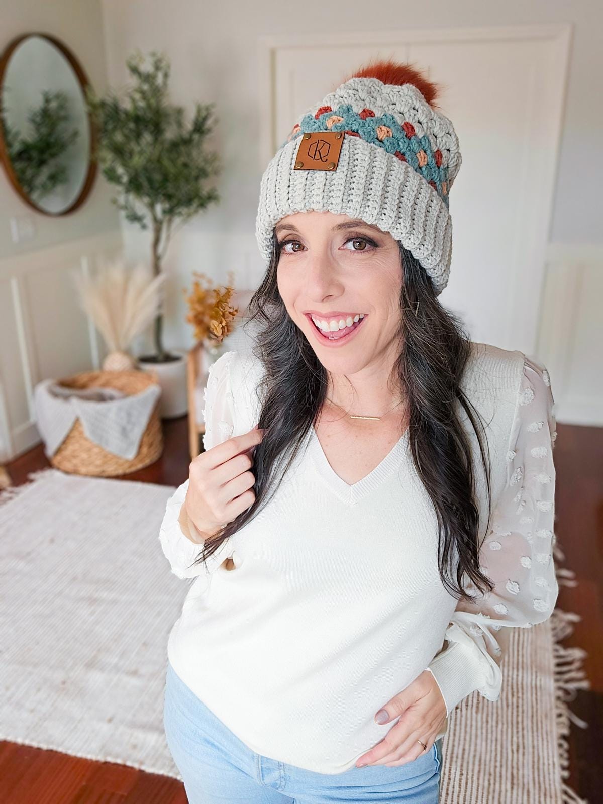 A woman wearing a crochet hat and a white blouse stands indoors, smiling, with unique jewellery accentuating her outfit. Home decor items like baskets and a rug add warmth to the background.