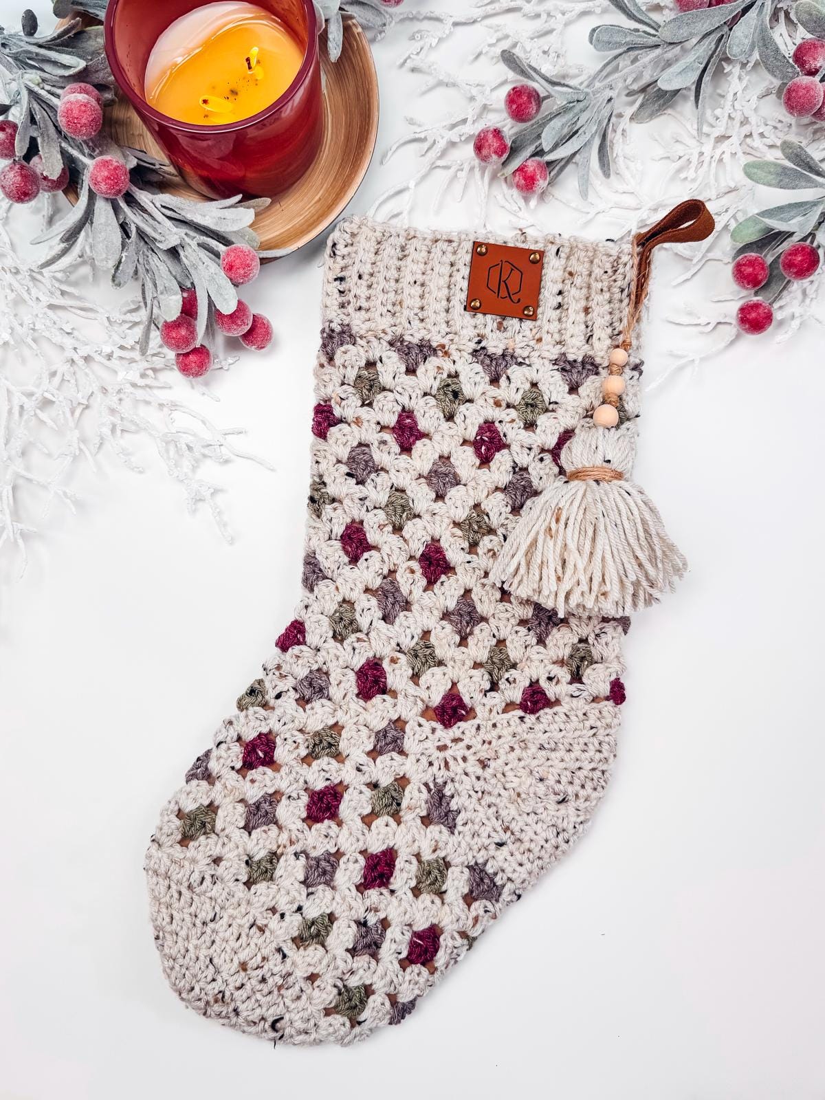 A crocheted stocking with a tassel is displayed on a white background. It features a multicolored pattern and is surrounded by faux snow-covered foliage and a lit candle, making it the perfect handmade gift.