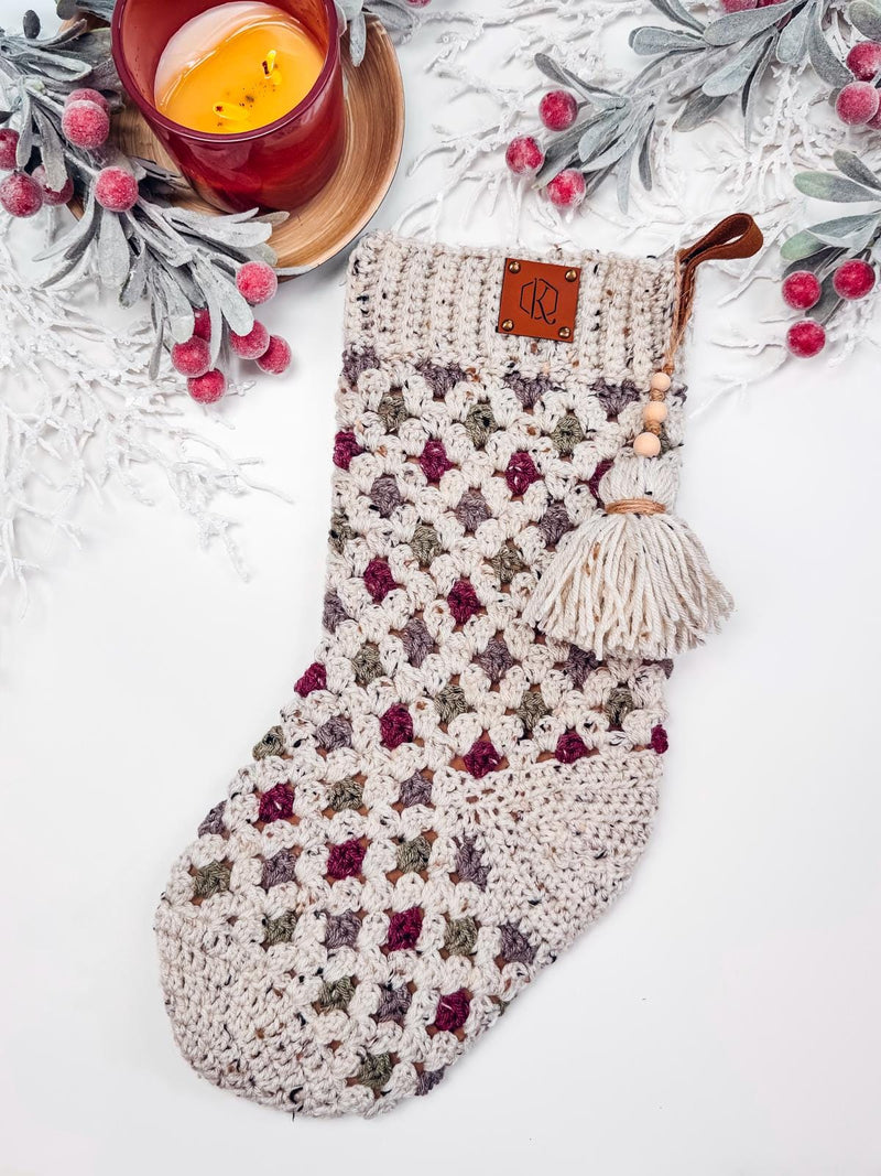 A crocheted stocking with a tassel is displayed on a white background. It features a multicolored pattern and is surrounded by faux snow-covered foliage and a lit candle, making it the perfect handmade gift.