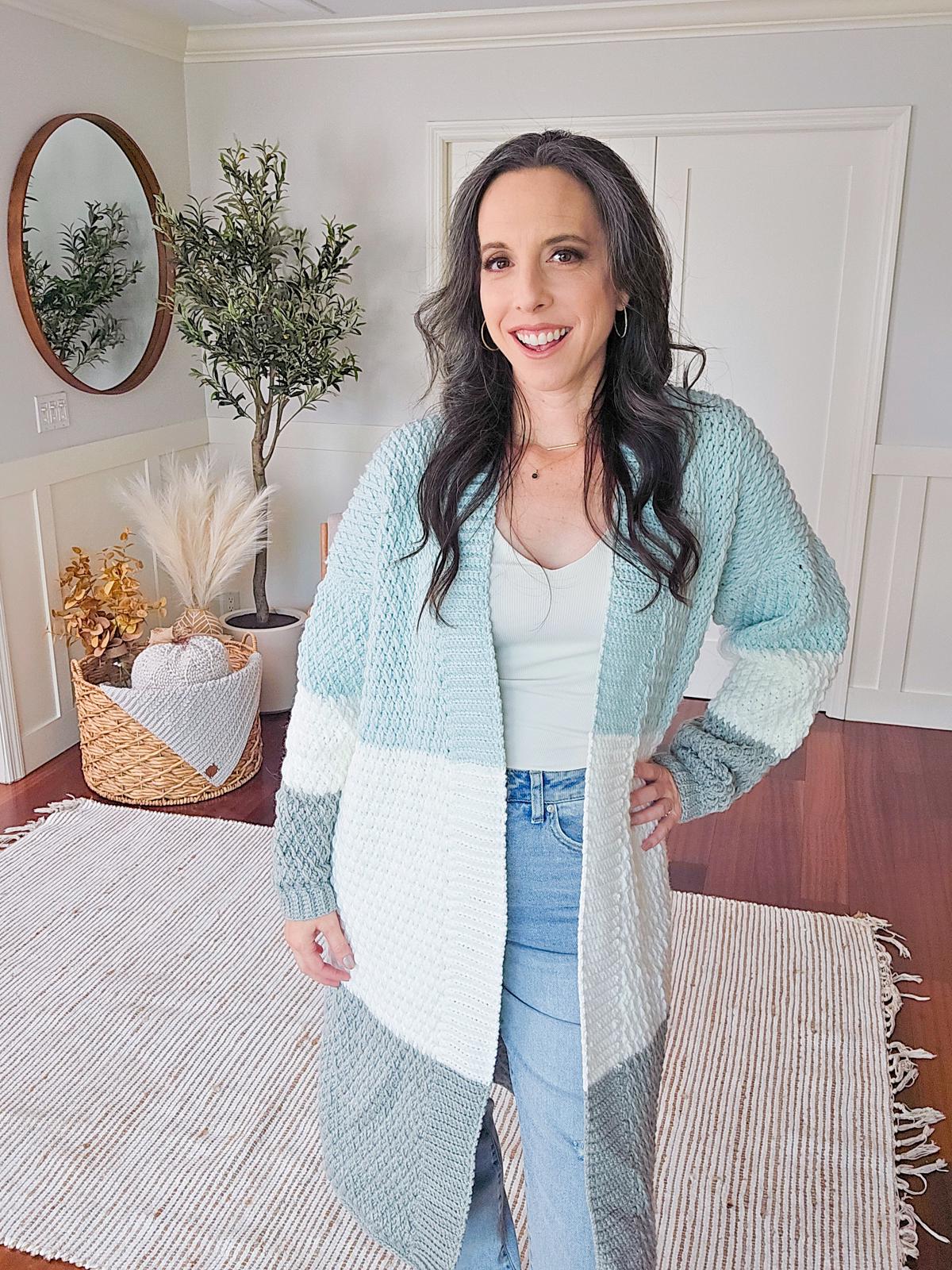 A person stands in a room with a woven rug, potted plant, and wall mirror. They wear a long, color-blocked cardigan reminiscent of vintage clothes, paired with a white top and jeans. A handmade gift lies beside them as they smile at the camera.