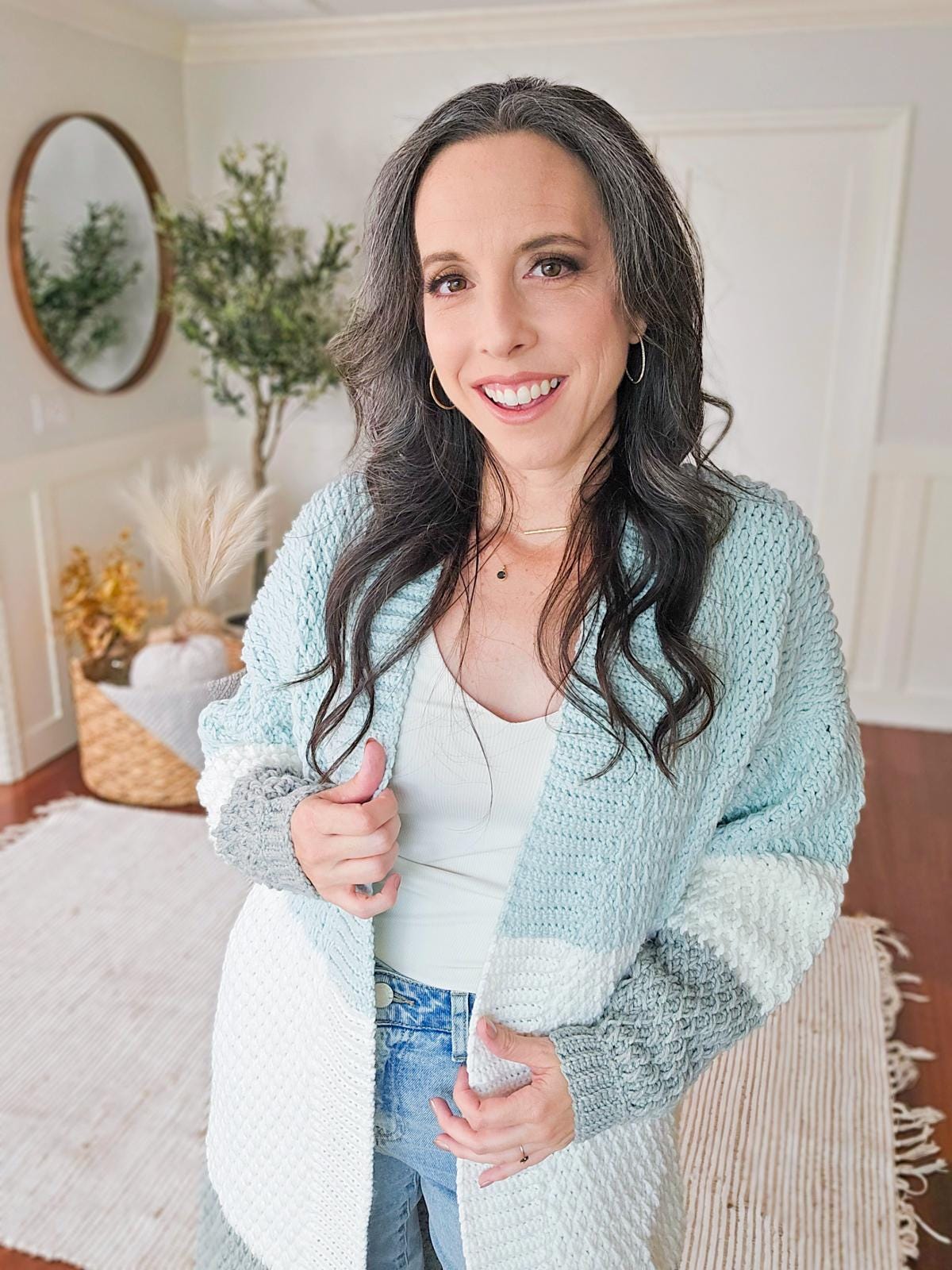 A person with long dark hair smiles at the camera, standing in a cozy room with a woven basket and a circular mirror. They're wearing vintage clothes—a light blue and gray knit cardigan and jeans.