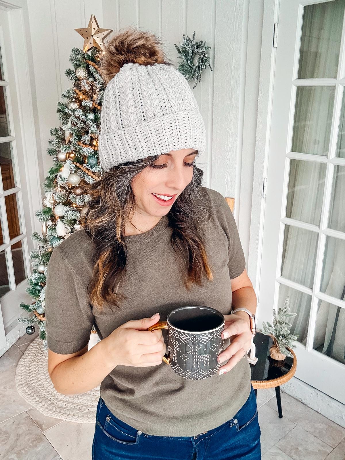 A person in a gray crochet hat, wearing vintage clothes, holds a mug while standing by a decorated Christmas tree indoors.