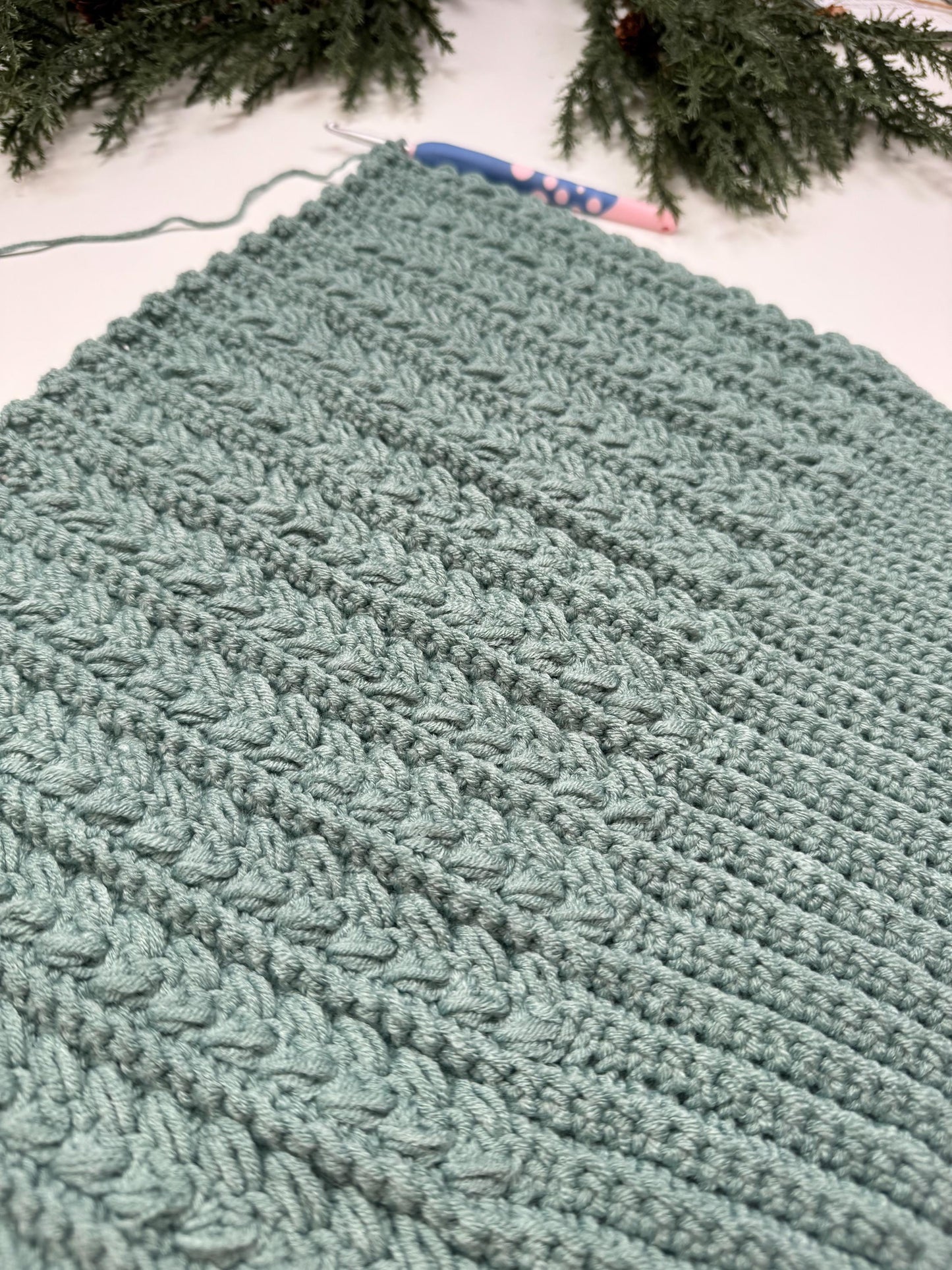 A teal crocheted blanket featuring textured patterns is laid out, with a crochet hook partially visible on top—an exquisite handmade gift that showcases the artistry and warmth of craftsmanship.