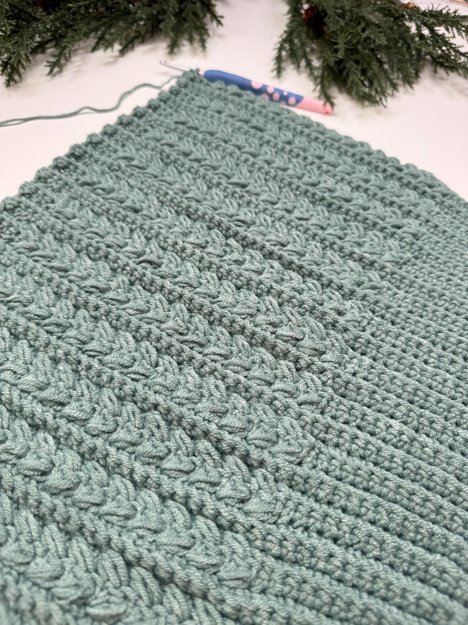 A teal crocheted blanket featuring textured patterns is laid out, with a crochet hook partially visible on top—an exquisite handmade gift that showcases the artistry and warmth of craftsmanship.