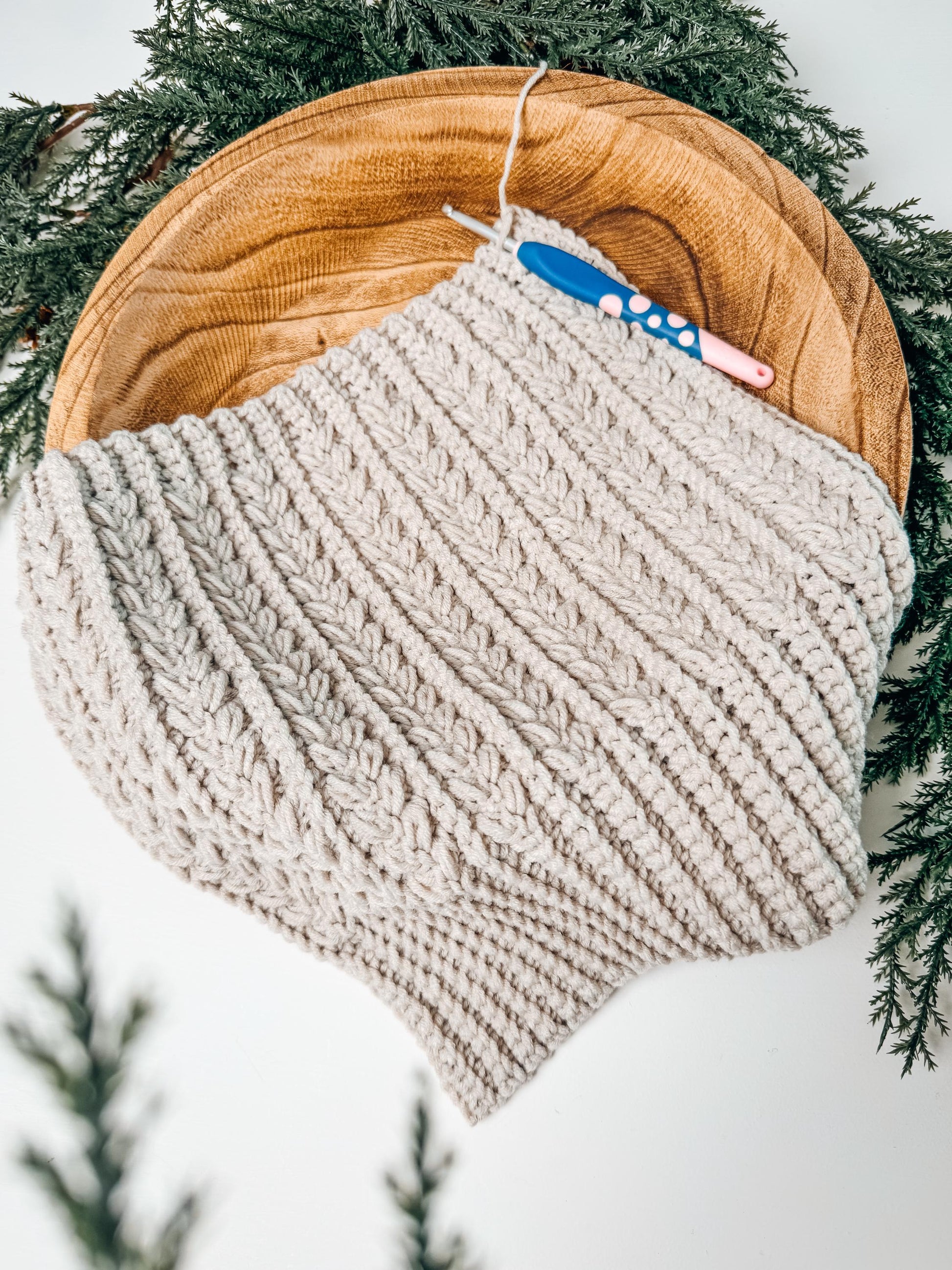 A knitted, cream-colored piece lies on a wooden plate with a crochet hook resting on top, surrounded by green foliage—an ideal handmade gift for those who appreciate cozy charm.
