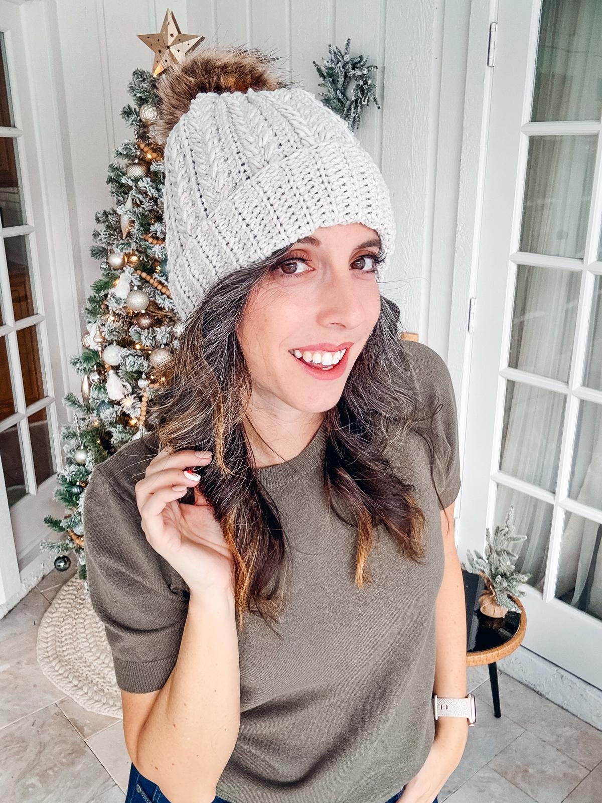 A woman wearing a gray knit hat smiles at the camera, showcasing her unique jewellery. There is a decorated Christmas tree in the background.