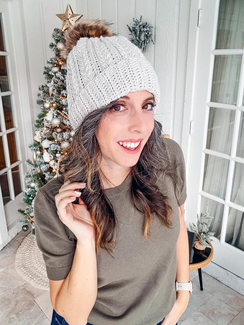 A woman wearing a gray knit hat smiles at the camera, showcasing her unique jewellery. There is a decorated Christmas tree in the background.