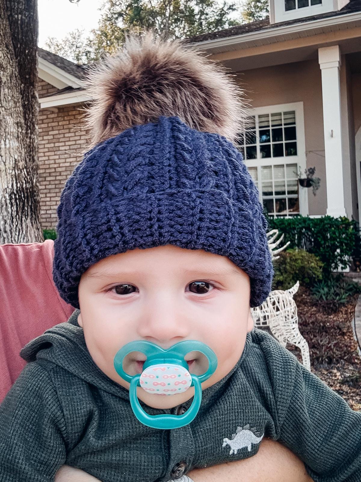 A baby with a pacifier, wearing a blue crochet hat with a fur pom-pom, sits outside in front of a house, resembling the charm of vintage clothes.