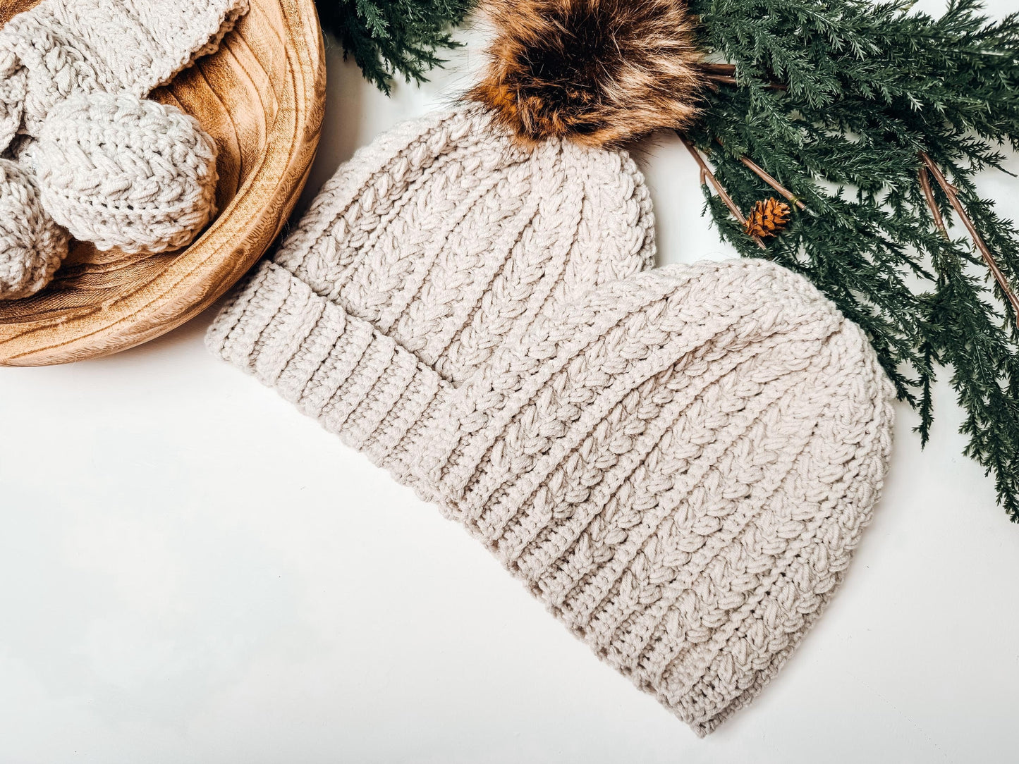 Two beige crochet hats with a fur pom-pom are placed alongside a wooden bowl brimming with knitted items and green pine branches, making for the perfect handmade gift.