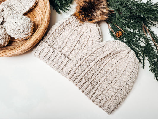 Two beige crochet hats with a fur pom-pom are placed alongside a wooden bowl brimming with knitted items and green pine branches, making for the perfect handmade gift.
