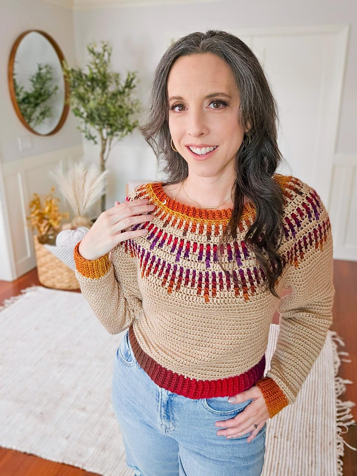 A person in a colorful crochet sweater smiles, standing on a vintage rug in a room with a mirror, unique jewelry glinting softly beside a thriving plant.
