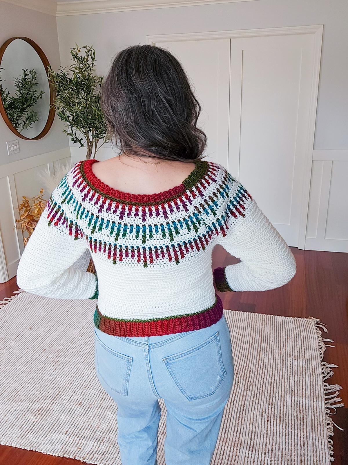 A person with long dark hair, wearing a white crochet sweater adorned with colorful patterns, stands facing a white wall featuring a mirror and plant.