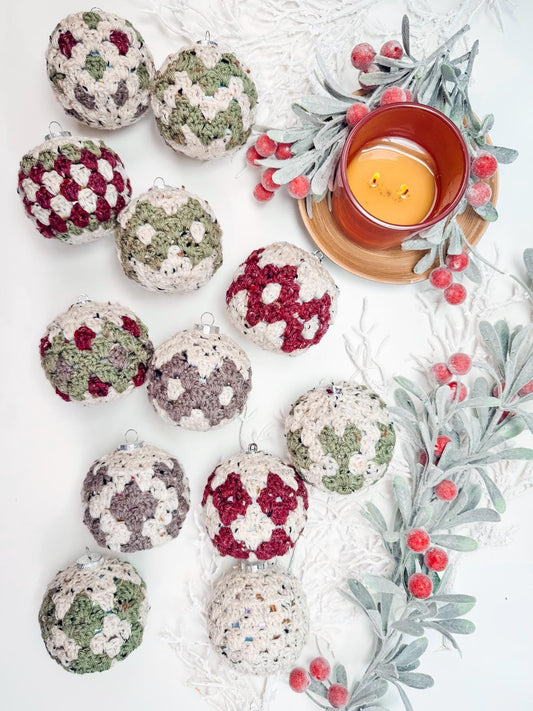 Handmade crochet ornaments in red, green, and white patterns are arranged beside a lit candle and frosted greenery with red berries on a white surface, creating a perfect setting for presenting a thoughtful handmade gift.