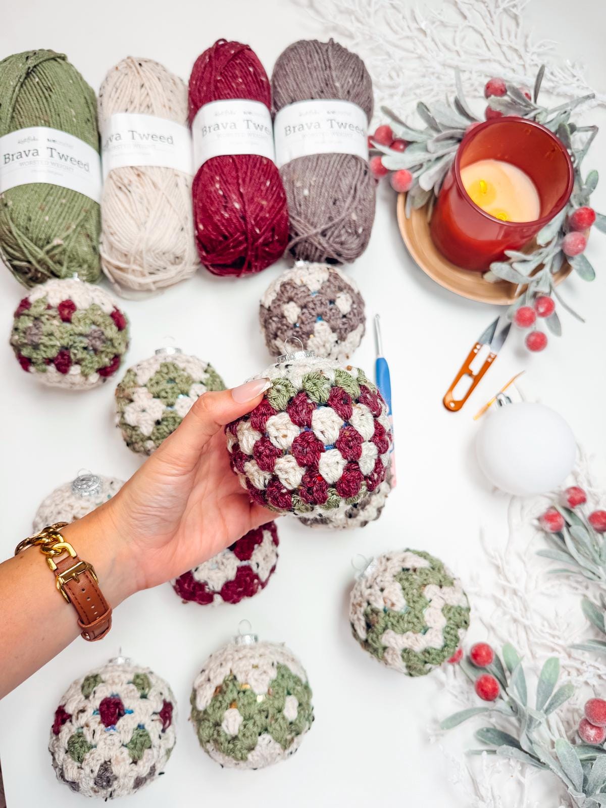 A hand holds a crocheted ornament above a table adorned with colorful yarns, a candle, and crochet tools — the perfect scene for crafting a handmade gift.
