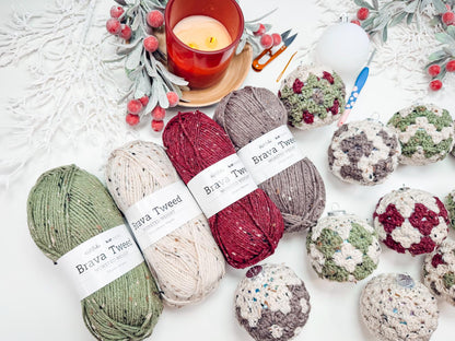 Yarn balls labeled "Brava Tweed" in green, cream, and maroon are arranged with crochet ornaments, a lit candle, scissors, and decorative greenery on a white surface—perfect for crafting a handmade gift.