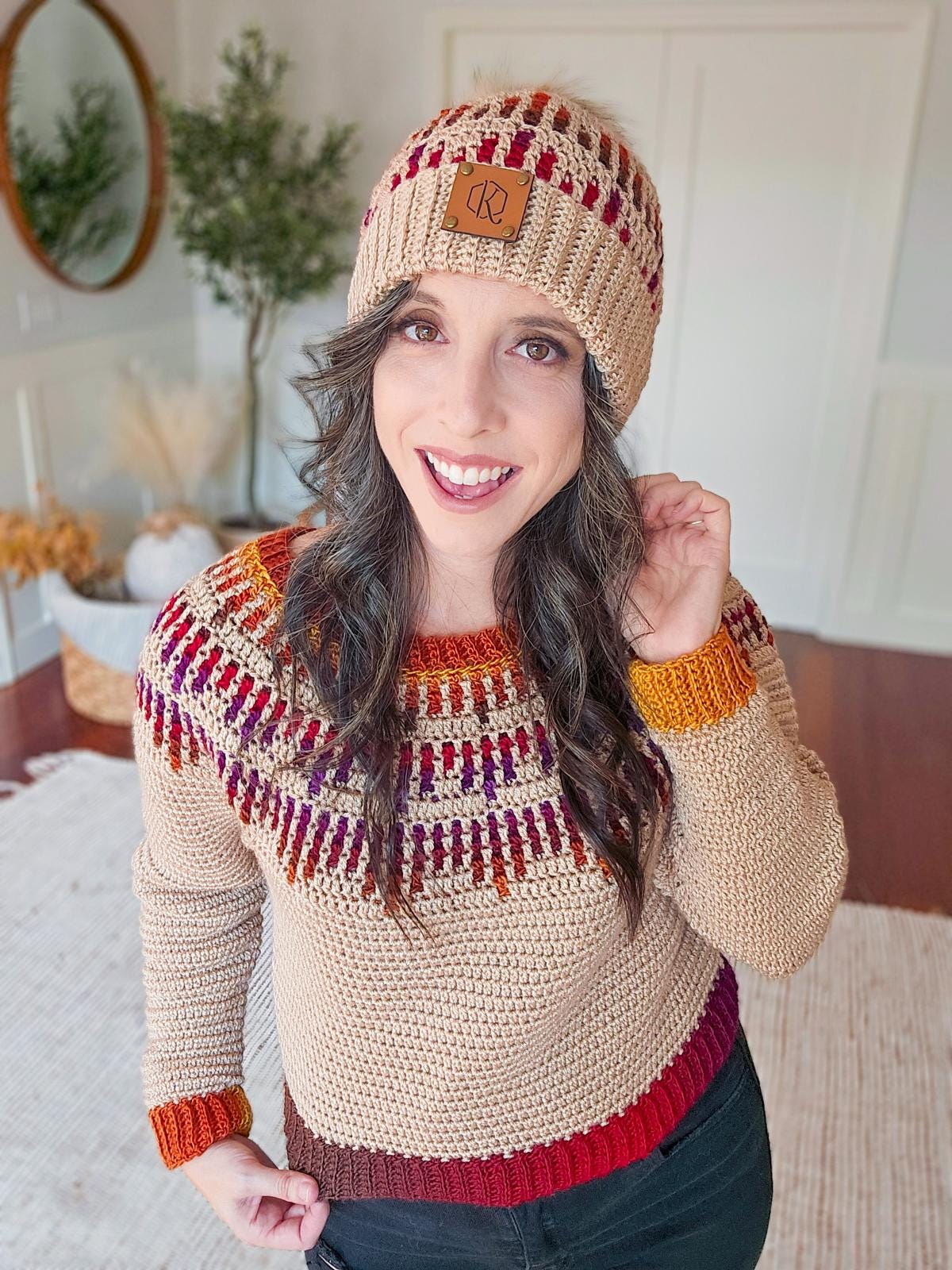 A person wearing a crocheted sweater and beanie with red and orange patterns stands smiling in a room adorned with a vintage mirror, alongside a plant.