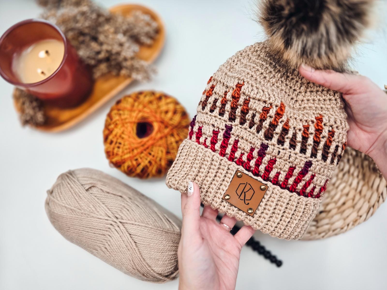 Hands holding a knitted hat with multicolored yarn and a pom-pom, near balls of colorful and white yarn, and a lit candle on the side—perfect as a handmade gift.
