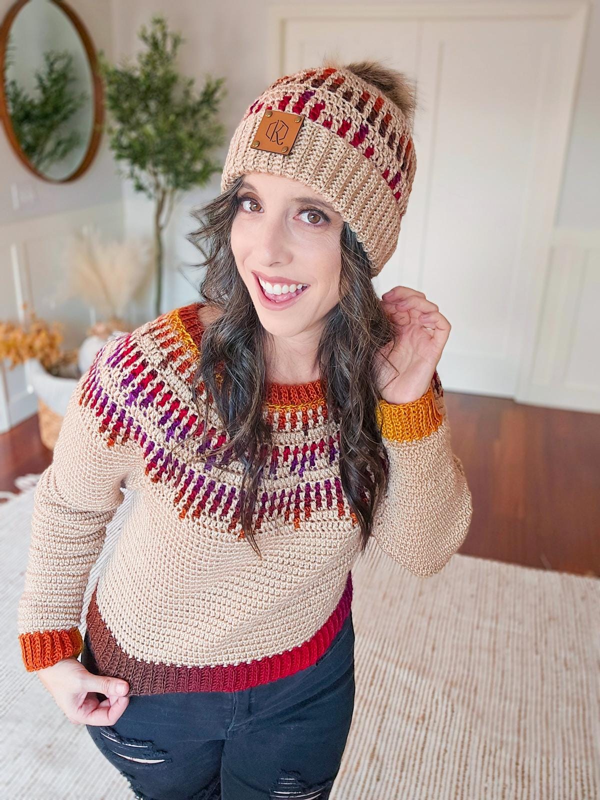 A person wearing a crocheted sweater and beanie with red and orange patterns stands smiling in a room adorned with a vintage mirror, alongside a plant.