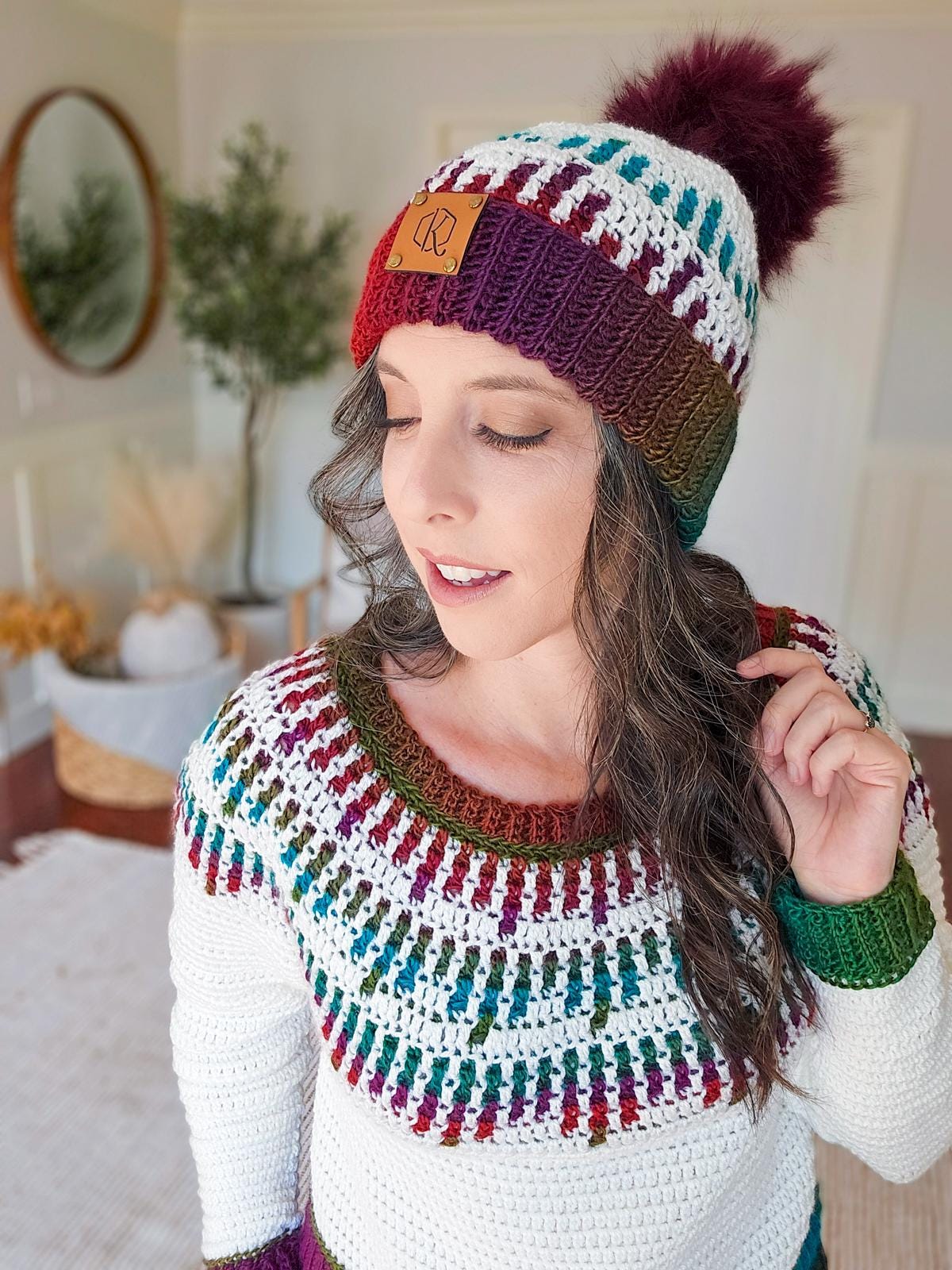 A person wearing a crocheted sweater and beanie with red and orange patterns stands smiling in a room adorned with a vintage mirror, alongside a plant.