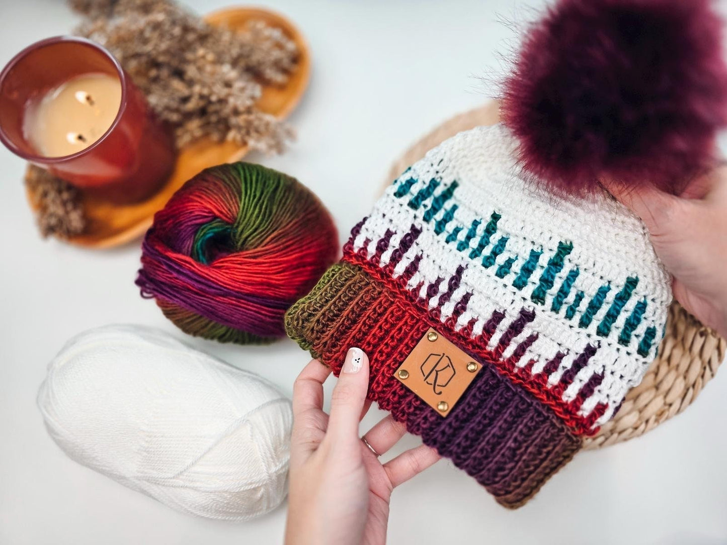 Hands holding a knitted hat with multicolored yarn and a pom-pom, near balls of colorful and white yarn, and a lit candle on the side—perfect as a handmade gift.