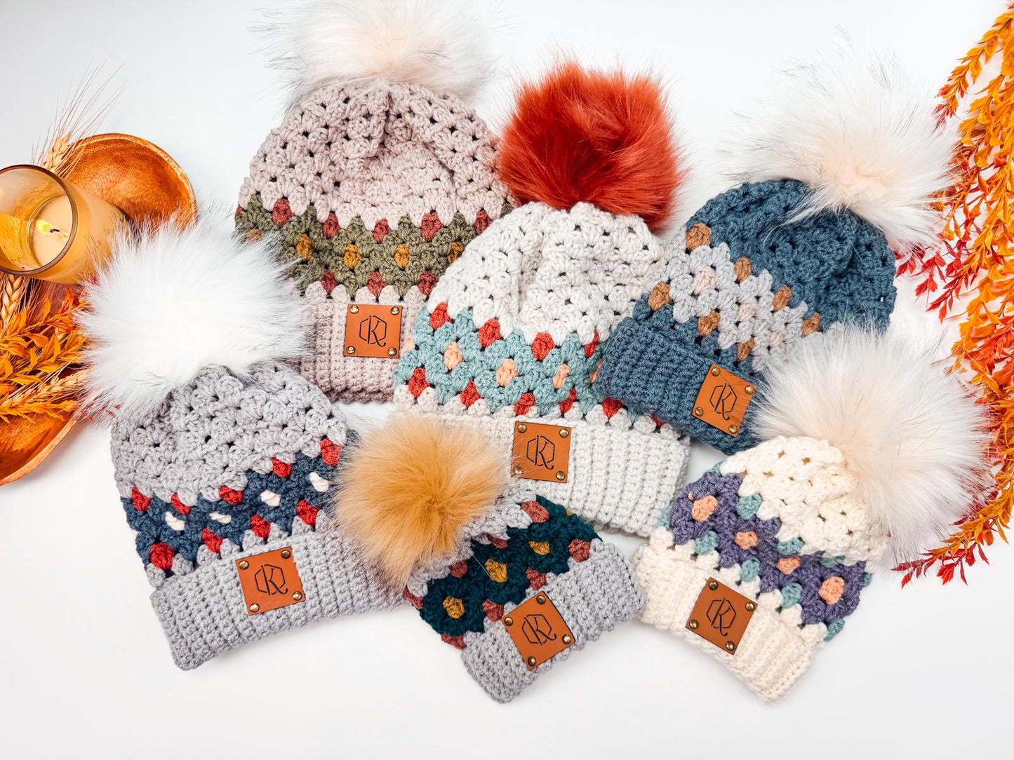 A collection of crocheted winter hats in various colors and patterns, each adorned with a fluffy pom-pom and a branded leather tag, displayed with autumn-themed decor. Perfect as a handmade gift for those who appreciate cozy elegance.