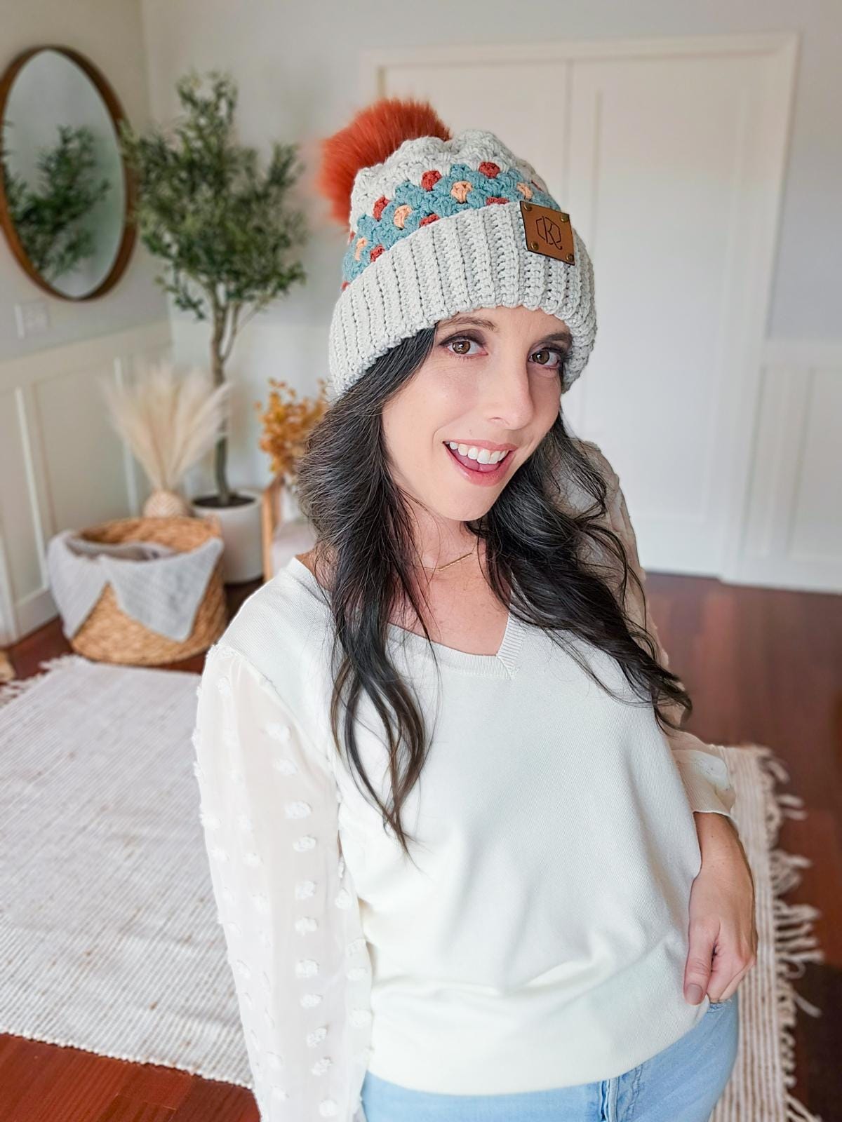 A woman in a cream sweater and knit hat with a pom-pom stands in a cozy, decorated room, adorned with vintage clothes, a mirror, basket, and dried plants.